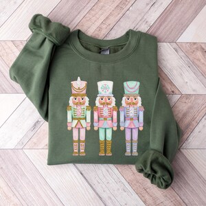 May include: An olive green sweatshirt featuring a colorful graphic of three nutcrackers. The nutcrackers are dressed in pastel colors with gold accents. The sweatshirt has long sleeves and a crew neck.
