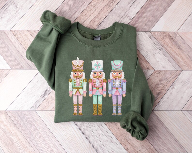 May include: An olive green sweatshirt featuring a colorful graphic of three nutcrackers. The nutcrackers are dressed in pastel colors with gold accents. The sweatshirt has long sleeves and a crew neck.