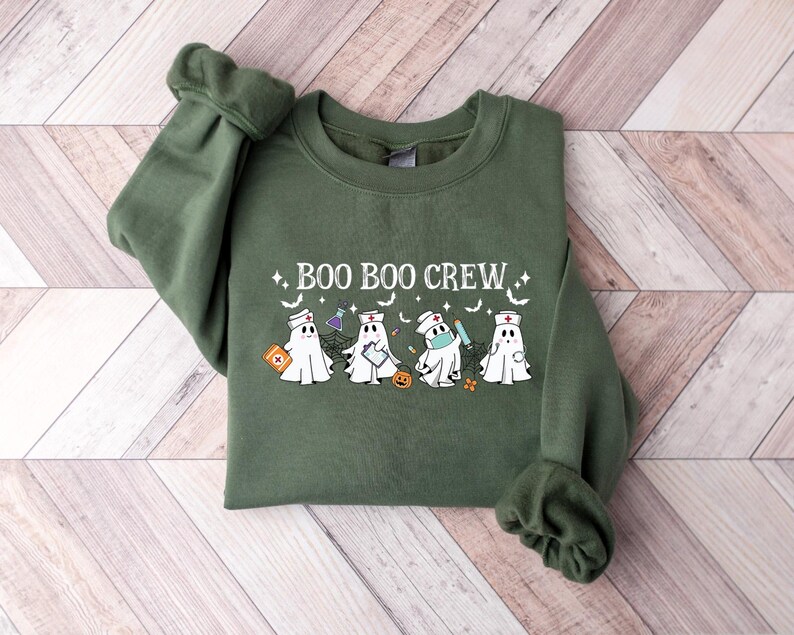 May include: An olive green sweatshirt featuring the text "BOO BOO CREW" and cartoon ghosts in medical attire. The ghosts are depicted with stethoscopes, face masks, and medical tools. The sweatshirt is made of a soft material and is a crew neck style.