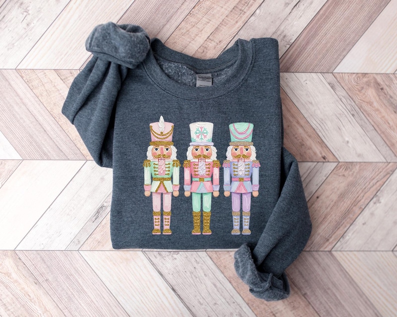 May include: A dark gray sweatshirt featuring three colorful nutcracker soldier designs. Each nutcracker has a different color scheme, including pink, green, and purple, with gold accents. The sweatshirt is folded on a light wood-grain surface.