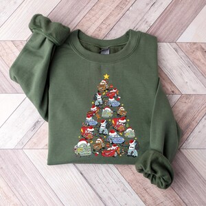 May include: An olive green sweatshirt featuring a Christmas tree design made of cartoon cars wearing Santa hats. The cars are in various colors, and a yellow star tops the tree. The sweatshirt has long sleeves and a crew neck.