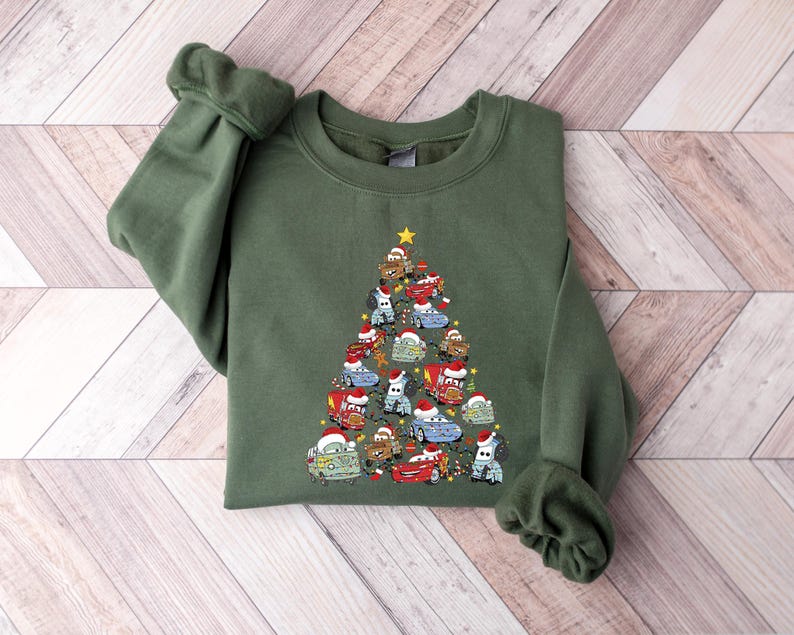 May include: An olive green sweatshirt featuring a Christmas tree design made of cartoon cars wearing Santa hats. The cars are in various colors, and a yellow star tops the tree. The sweatshirt has long sleeves and a crew neck.