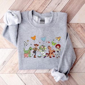 May include: A light gray sweatshirt featuring a colorful graphic of characters from the animated movie Toy Story. The design includes Woody, Buzz Lightyear, and other characters, along with star and balloon illustrations.