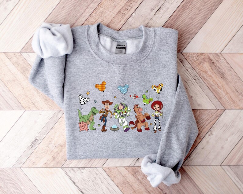 May include: A light gray sweatshirt featuring a colorful graphic of characters from the animated movie Toy Story. The design includes Woody, Buzz Lightyear, and other characters, along with star and balloon illustrations.