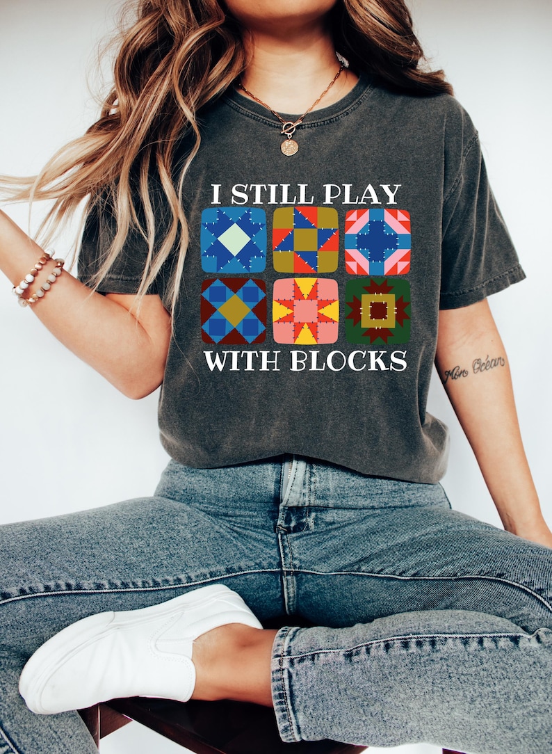 May include: Dark gray t-shirt with the text "I STILL PLAY WITH BLOCKS" above a grid of colorful quilt block designs. The shirt is paired with blue jeans and white sneakers.
