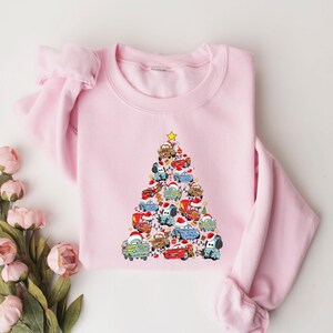 May include: A light pink sweatshirt featuring a Christmas tree design made of cartoon cars. The cars are decorated with Santa hats and other festive elements. A yellow star tops the tree. The sweatshirt is a crew neck style.