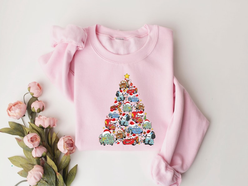 May include: A light pink sweatshirt featuring a Christmas tree design made of cartoon cars. The cars are decorated with Santa hats and other festive elements. A yellow star tops the tree. The sweatshirt is a crew neck style.