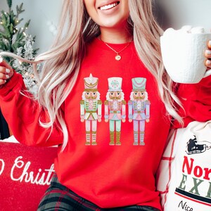 May include: A red sweatshirt featuring three colorful nutcracker soldier illustrations. The nutcrackers are in pastel colors with gold accents. The word "Christ" is visible on a pillow. A white mug is held in the hand.