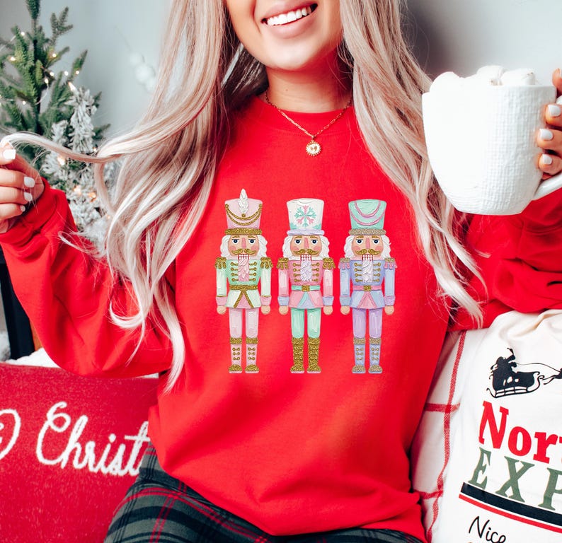 May include: A red sweatshirt featuring three colorful nutcracker soldier illustrations. The nutcrackers are in pastel colors with gold accents. The word "Christ" is visible on a pillow. A white mug is held in the hand.