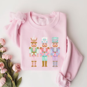May include: A light pink sweatshirt featuring three colorful nutcracker figures. Each nutcracker has a different hat and uniform, with colors including mint green, pink, and blue. The sweatshirt is made of a soft material.