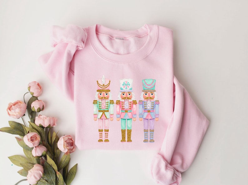 May include: A light pink sweatshirt featuring three colorful nutcracker figures. Each nutcracker has a different hat and uniform, with colors including mint green, pink, and blue. The sweatshirt is made of a soft material.