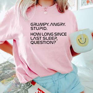 May include: A light pink t-shirt with the text "GRUMPY. ANGRY. STUPID. HOW LONG SINCE LAST SLEEP, QUESTION?" printed in black. The person is holding a white mug with a yellow smiley face.