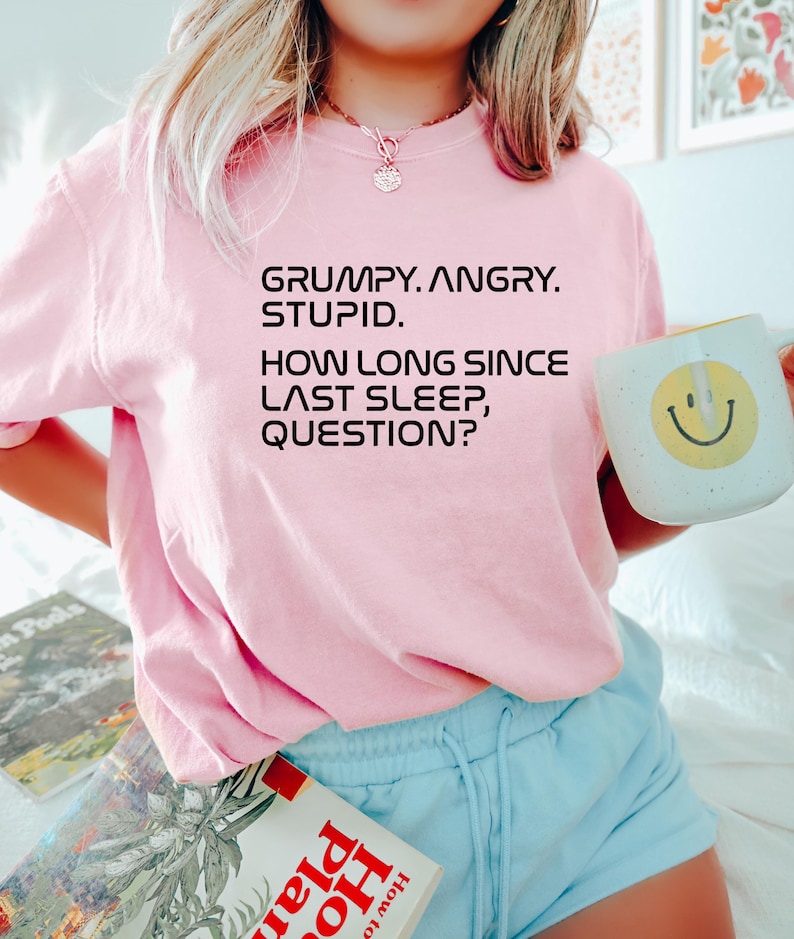 May include: A light pink t-shirt with the text "GRUMPY. ANGRY. STUPID. HOW LONG SINCE LAST SLEEP, QUESTION?" printed in black. The person is holding a white mug with a yellow smiley face.