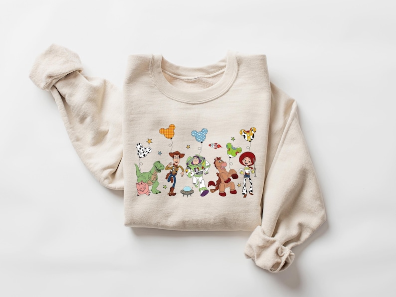 May include: A beige sweatshirt featuring a colorful graphic of animated characters and balloons. The design includes a dinosaur, a pig, a cowboy, a space ranger, a horse, and a cowgirl. The balloons have various patterns and shapes.