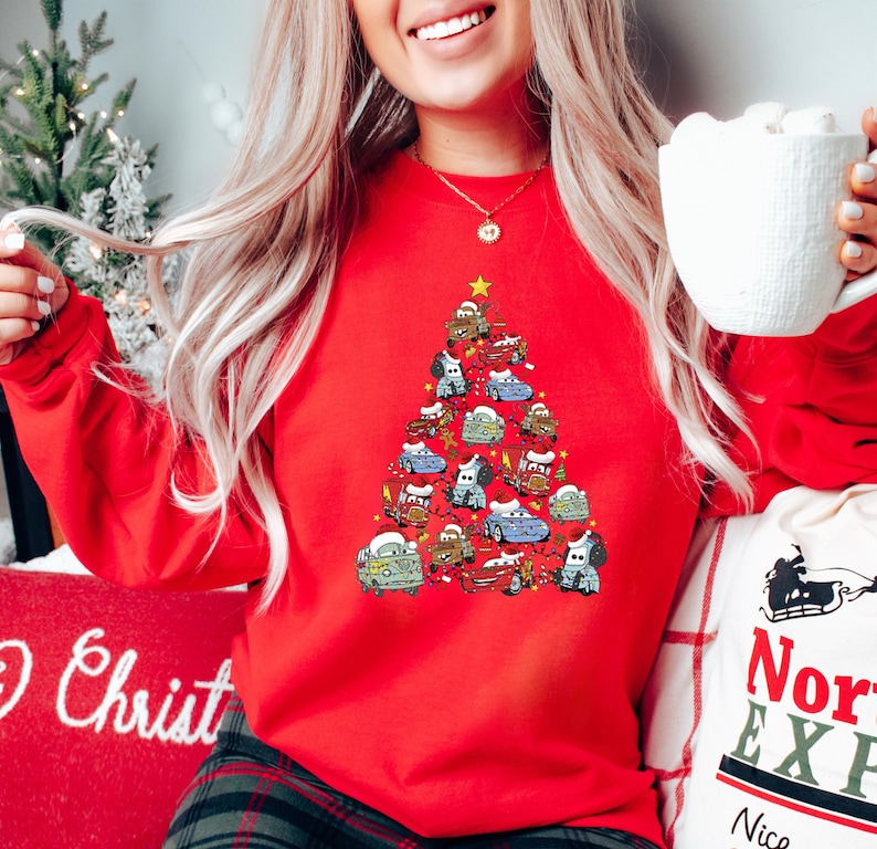 May include: A red sweatshirt featuring a Christmas tree design made of cartoon cars. The cars are in various colors and decorated with festive elements. A gold star tops the tree. The sweatshirt is part of a holiday collection.
