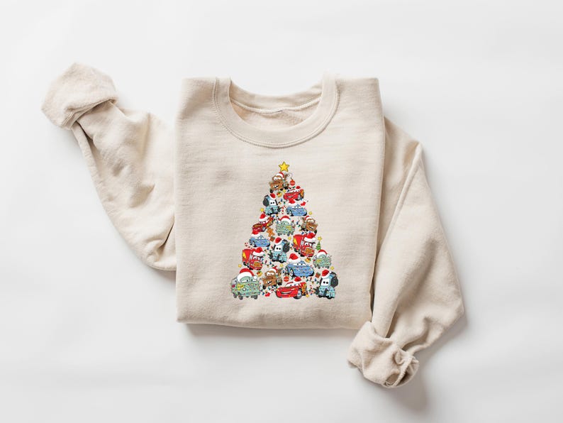 May include: A beige sweatshirt featuring a Christmas tree design made of various cartoon cars. The cars are in different colors and are wearing Santa hats. A yellow star tops the tree. The sweatshirt is folded on a white surface.