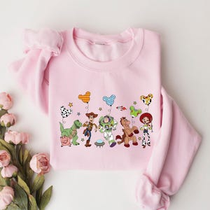 May include: A light pink sweatshirt featuring a colorful graphic of animated characters holding balloons. The design includes a green dinosaur, a cowboy, a space ranger, a horse, and a cowgirl. The sweatshirt has long sleeves.