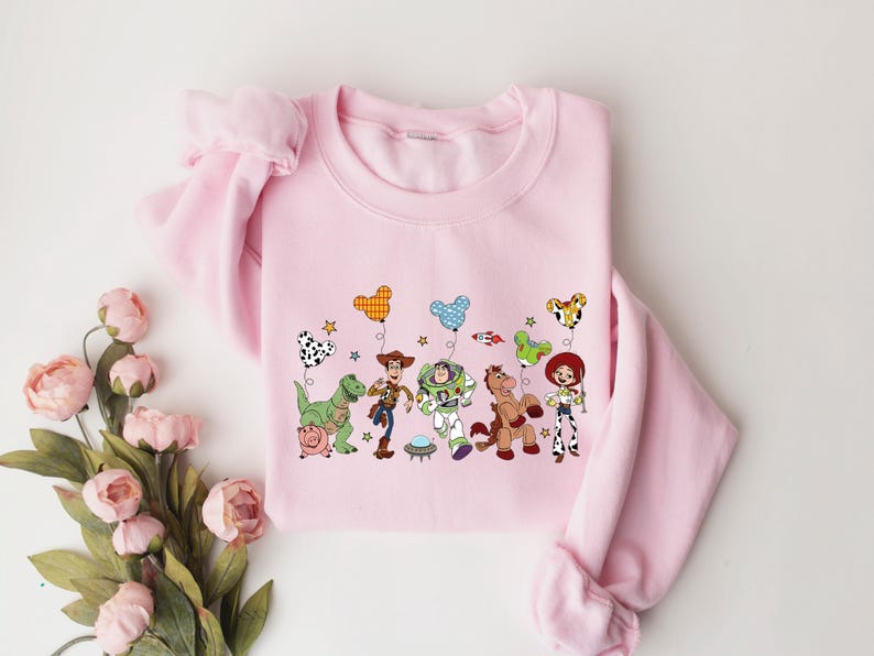 May include: A light pink sweatshirt featuring a colorful graphic of animated characters holding balloons. The design includes a green dinosaur, a cowboy, a space ranger, a horse, and a cowgirl. The sweatshirt has long sleeves.