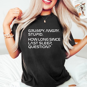 May include: Black t-shirt with the text "GRUMPY. ANGRY. STUPID. HOW LONG SINCE LAST SLEEP, QUESTION?" in white. The shirt is a crew neck style.