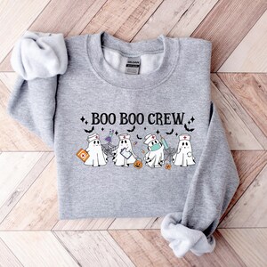 May include: A grey sweatshirt featuring the words "BOO BOO CREW" and ghost illustrations dressed as nurses, some wearing face masks, with medical items. The design includes bats, stars, and a pumpkin.