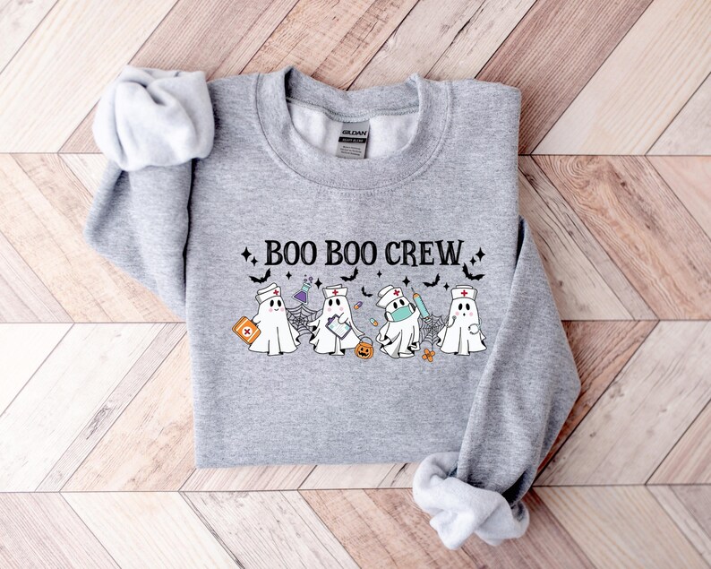 May include: A grey sweatshirt featuring the words "BOO BOO CREW" and ghost illustrations dressed as nurses, some wearing face masks, with medical items. The design includes bats, stars, and a pumpkin.
