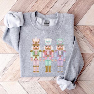 May include: A light gray sweatshirt featuring three colorful nutcracker soldier illustrations. Each nutcracker is adorned with a different pastel color scheme, including pink, green, and blue, with gold accents. The sweatshirt is folded on a wooden surface.