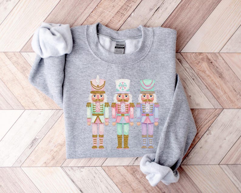May include: A light gray sweatshirt featuring three colorful nutcracker soldier illustrations. Each nutcracker is adorned with a different pastel color scheme, including pink, green, and blue, with gold accents. The sweatshirt is folded on a wooden surface.