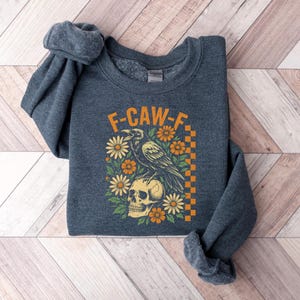 May include: A dark gray sweatshirt with the text "F-CAW-F" in orange. The graphic features a raven perched on a skull, surrounded by orange and yellow flowers. A checkered orange and yellow pattern is on the right side.