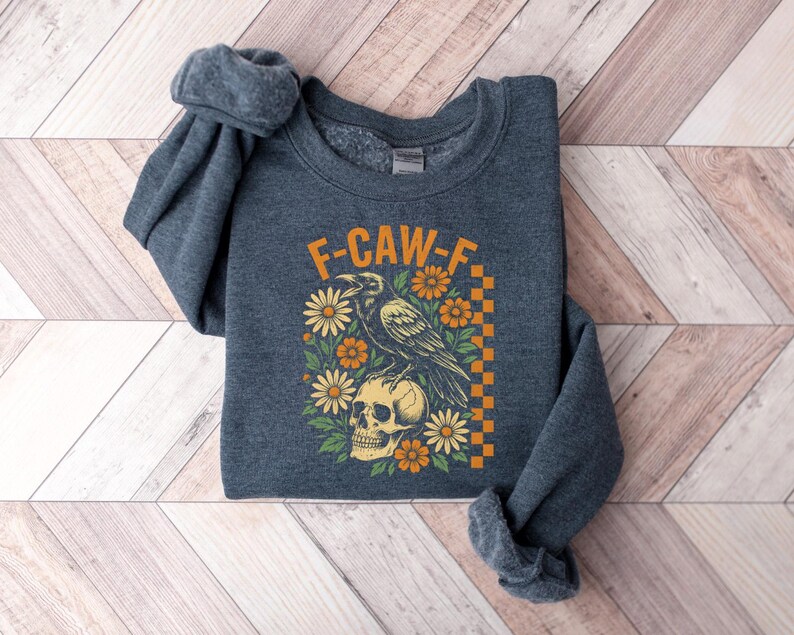 May include: A dark gray sweatshirt with the text "F-CAW-F" in orange. The graphic features a raven perched on a skull, surrounded by orange and yellow flowers. A checkered orange and yellow pattern is on the right side.