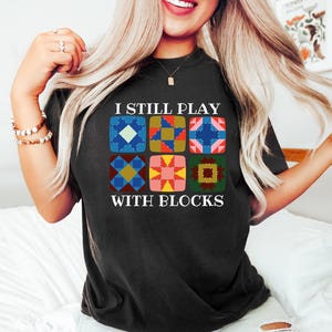 May include: Black t-shirt with the text "I Still Play With Blocks" and a colorful quilt block design. The design features six different quilt block patterns in various colors, including blue, pink, yellow, and green.