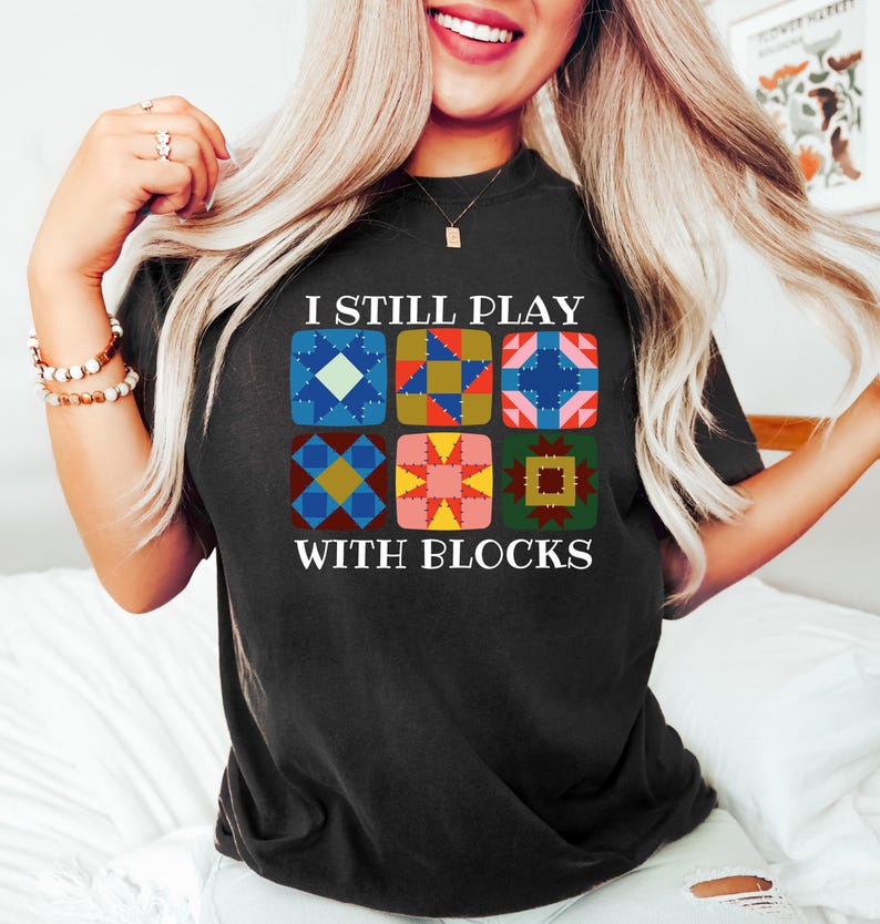 May include: Black t-shirt with the text "I Still Play With Blocks" and a colorful quilt block design. The design features six different quilt block patterns in various colors, including blue, pink, yellow, and green.