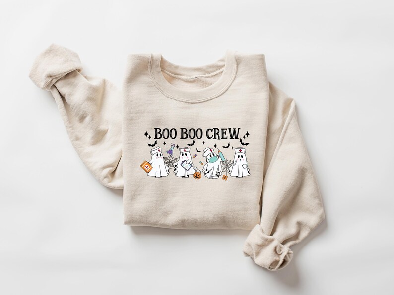 May include: A cream-coloured sweatshirt featuring the words "BOO BOO CREW" and illustrations of ghosts in medical attire. The ghosts are depicted with nurse hats, masks, and medical equipment. Halloween-themed clothing.