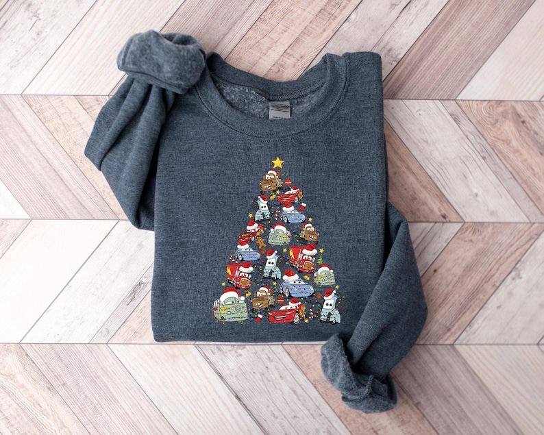 May include: A dark gray sweatshirt featuring a Christmas tree design made of cartoon cars. The cars are decorated with Santa hats and a gold star at the top. The sweatshirt is made of a soft material.