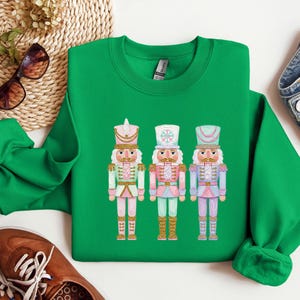 May include: A green sweatshirt featuring three colorful nutcracker illustrations. Each nutcracker is dressed in a different pastel color scheme, with gold accents and detailed hats. The design is centered on the front of the sweatshirt.