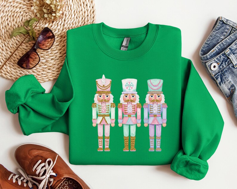 May include: A green sweatshirt featuring three colorful nutcracker illustrations. Each nutcracker is dressed in a different pastel color scheme, with gold accents and detailed hats. The design is centered on the front of the sweatshirt.
