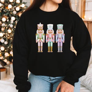 May include: Black crewneck sweatshirt featuring a colorful graphic of three nutcracker figures. The nutcrackers are in pastel colors of pink, green, and blue, with gold accents. The sweatshirt is a casual style, suitable for the holiday season.