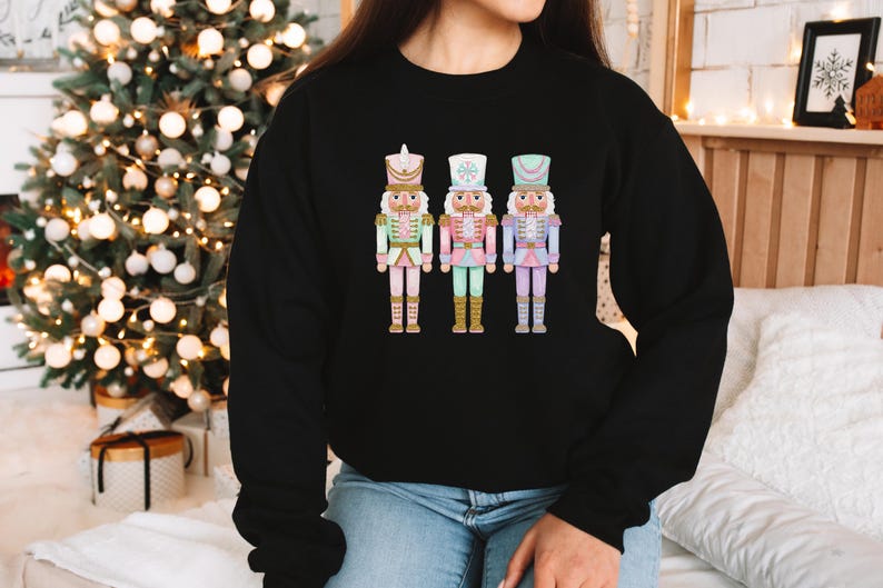 May include: Black crewneck sweatshirt featuring a colorful graphic of three nutcracker figures. The nutcrackers are in pastel colors of pink, green, and blue, with gold accents. The sweatshirt is a casual style, suitable for the holiday season.