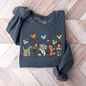 May include: A dark gray sweatshirt featuring a colorful graphic of characters from a popular animated movie. The design includes a dinosaur, a cowboy, a space ranger, a horse, and a cowgirl, along with other cartoon elements.