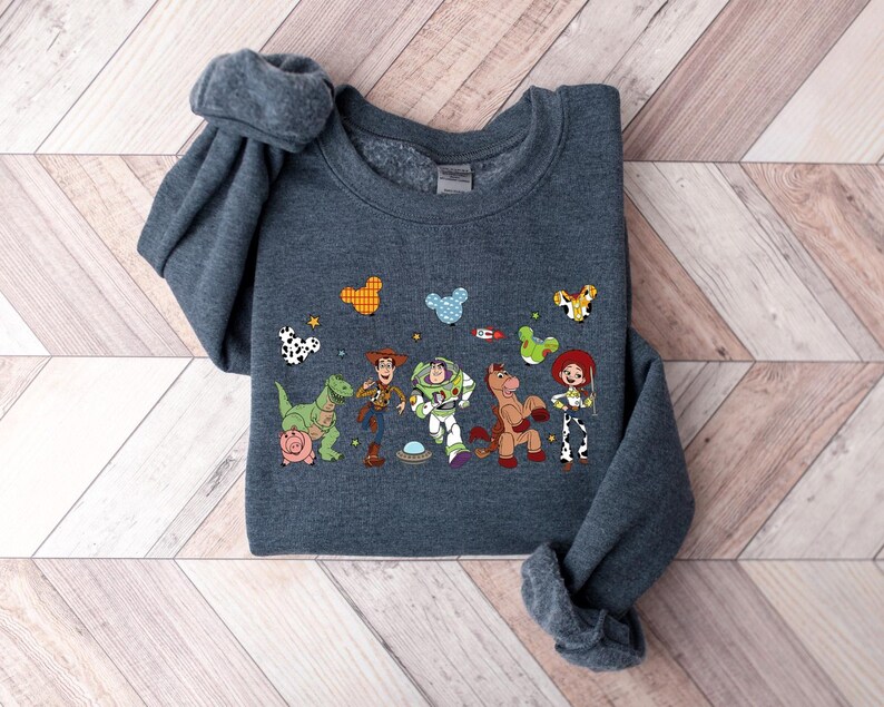 May include: A dark gray sweatshirt featuring a colorful graphic of characters from a popular animated movie. The design includes a dinosaur, a cowboy, a space ranger, a horse, and a cowgirl, along with other cartoon elements.
