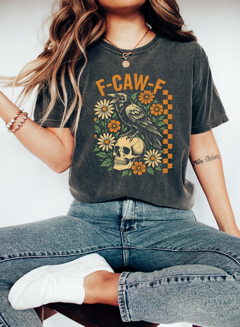 May include: Dark gray t-shirt featuring a graphic design with the text "F-CAW-F". The design includes a raven perched on a skull, surrounded by orange and white flowers. The shirt is worn with blue jeans and white shoes.
