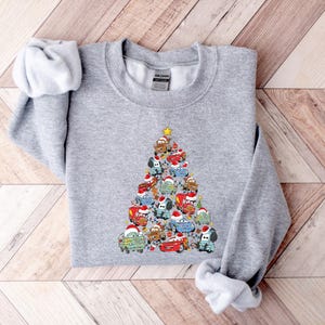 May include: A light gray sweatshirt featuring a Christmas tree design made of cartoon cars wearing Santa hats. The cars are various colors, and a yellow star tops the tree. The sweatshirt is made of a soft material.