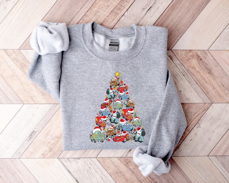 May include: A light gray sweatshirt featuring a Christmas tree design made of cartoon cars wearing Santa hats. The cars are various colors, and a yellow star tops the tree. The sweatshirt is made of a soft material.