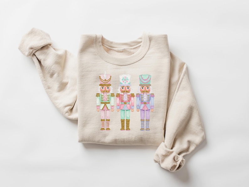 May include: Beige sweatshirt featuring a watercolor design of three toy nutcracker soldiers in pastel colors. The nutcrackers have gold accents and are dressed in pink, green, and blue uniforms. A cozy, long-sleeved top.