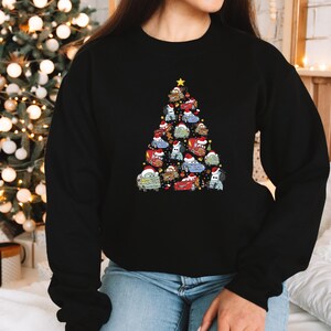 May include: Black sweatshirt featuring a Christmas tree design made of cartoon cars wearing Santa hats. The tree is topped with a gold star. The sweatshirt is worn by a person in blue jeans.