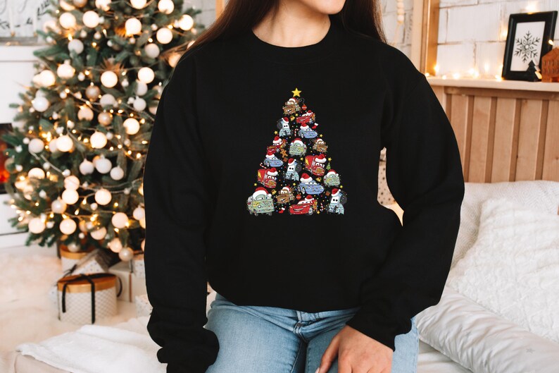 May include: Black sweatshirt featuring a Christmas tree design made of cartoon cars wearing Santa hats. The tree is topped with a gold star. The sweatshirt is worn by a person in blue jeans.