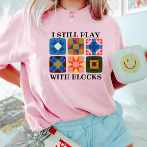 May include: A light pink sweatshirt with the text "I STILL PLAY WITH BLOCKS" and a graphic of six colorful quilt block designs. The person is holding a white mug with a yellow smiley face.