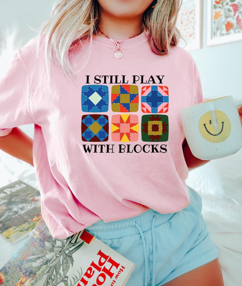 May include: A light pink sweatshirt with the text "I STILL PLAY WITH BLOCKS" and a graphic of six colorful quilt block designs. The person is holding a white mug with a yellow smiley face.