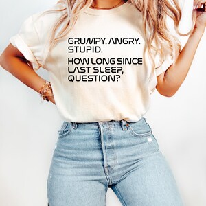 May include: Cream-colored t-shirt with the text "GRUMPY. ANGRY. STUPID. HOW LONG SINCE LAST SLEEP, QUESTION?" in black. The shirt is paired with light blue denim jeans.
