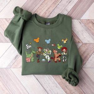 May include: Olive green sweatshirt featuring a colorful graphic of characters from a popular animated movie. The design includes a dinosaur, a pig, a cowboy, a space ranger, a horse, and a cowgirl. The sweatshirt has long sleeves and a crew neck.