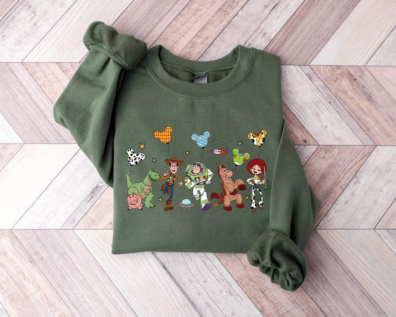 May include: Olive green sweatshirt featuring a colorful graphic of characters from a popular animated movie. The design includes a dinosaur, a pig, a cowboy, a space ranger, a horse, and a cowgirl. The sweatshirt has long sleeves and a crew neck.